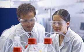 Two individuals in white lab coats are engaged in a discussion in a laboratory setting. They are surrounded by laboratory equipment, including flasks and bottles, suggesting a focus on scientific research or experimentation.