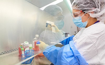 A person in protective clothing, including a hairnet and face mask, working in a laboratory with various bottles of chemicals on the countertop. Background with a clean, organized workspace, including a laptop and scientific equipment.