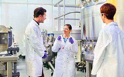 Three scientists are engaged in a discussion within a laboratory, surrounded by stainless steel equipment.