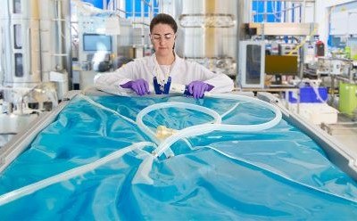 A person in a lab coat and purple gloves stands behind a large, transparent blue plastic container filled with liquid. Tubing is connected to the container, and the background shows various laboratory equipment and machinery. A person in a lab coat and purple gloves stands behind a large, transparent blue plastic container filled with liquid. Tubing is connected to the container, and the background shows various laboratory equipment and machinery.