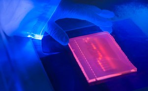 A close-up view of a blue-lit laboratory setting. The focus is on a small, glowing rectangular object that emits a pinkish-red light. The object is placed on top of another piece of equipment with a dark surface.
