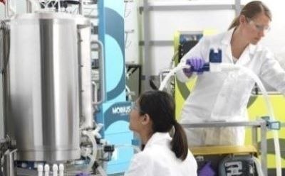 Bioprocessing cell culture media (CCM) The image depicts a laboratory scene with two professionals working on a large stainless steel equipment. They are both wearing white lab coats and gloves, suggesting a sterile environment. One person is standing and appears to be handling something with purple gloves, while the other is seated. The equipment is connected to various tubes and wires, indicating its complex functionality.