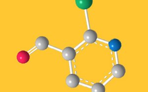 A 3D molecular structure illustration on a yellow background. The model shows a ring structure with gray spheres representing carbon atoms, connected by white bonds. Various colored spheres represent different atoms: a red sphere (possibly oxygen), a green sphere, and a blue sphere. The arrangement highlights the molecular connectivity and geometry.