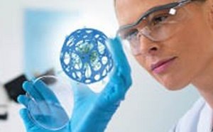 A scientist in a lab coat and blue gloves is examining a detailed blue 3D model of a molecular structure with blurred laboratory equipment in the background.