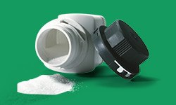 A white, overturned supplement bottle with a black cap detached and lying next to it. A pile of white powder is spilled from the bottle onto a green surface.