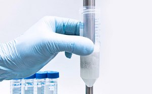 A close-up image of a gloved hand holding a clear centrifuge tube containing a white precipitate at the bottom. The tube is marked with measurement lines, and there are additional tubes in the background, all against a clean, light blue backdrop. This image is relevant as it depicts a common laboratory procedure for separating substances based on density, which is essential in many scientific research and diagnostic processes.