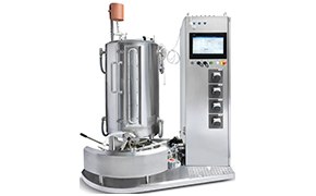 A machine with a vertical cylindrical chamber and various attached pipes, gauges, and valves, accompanied by an electronic control panel with a screen to the right side.