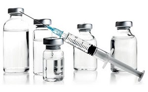 A medical syringe with a needle, partially filled liquid, is positioned diagonally in front of five vials of various sizes with metallic caps against a white background.