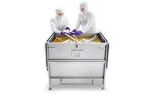 Two people in protective lab attire are interacting with the contents of a large metal container. The container has latches on the side, indicating it may has a lid. 
