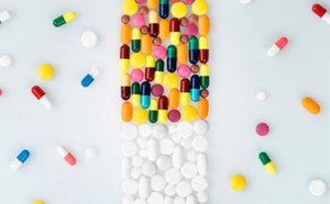 The image depicts an array of colorful pills and capsules artistically arranged on a white surface to resemble the shape of a pill bottle, with the body composed of multi-colored capsules and the cap made of white round tablets, surrounded by additional loose pills.