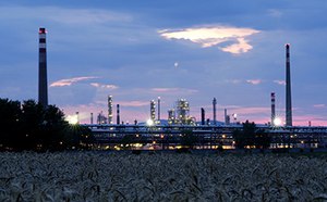 The image depicts an industrial plant at twilight, with tall chimneys and illuminated structures set against a cloudy sky, viewed over a field of crops.