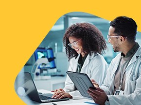 Two laboratory technicians in white lab coats are engaged in a discussion while reviewing information on a laptop and a tablet. The background features laboratory equipment, including a microscope, indicating a collaborative scientific environment.