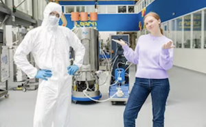 Two scientists, standing in a lab, a male in bunny suit, a female wearing casual clothes