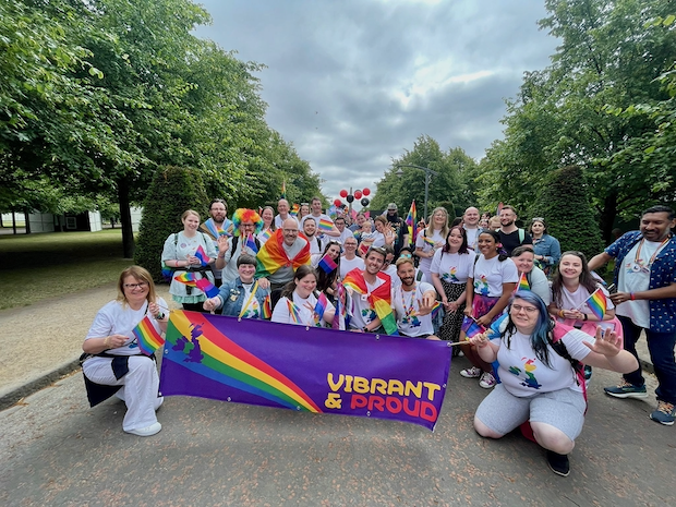 Cork Pride 2022 약 30명의 직원이 레인보우 깃발과 함께 '힘차고 당당하게(Vibrant and Proud)'라고 쓰인 현수막을 들고 있습니다. 그들은 레인보우 깃발을 들고 환하게 웃고 있습니다.