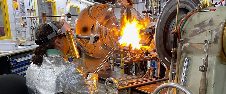 Glassblower works on flask 둥근 바닥 플라스크가 선반 위에 놓여 있고 유리 직공이 플라스크의 목 부분에 불꽃을 겨냥합니다. 유리 직공은 특수 제작한 겉옷, 장갑, 안면 보호대 등 보호복을 착용합니다.