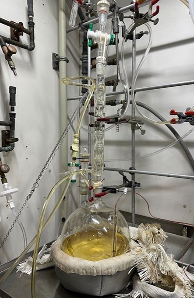 Round-bottom flask 튜브가 있는 둥근 바닥 플라스크가 실험실 벤치에 놓여 있습니다. 내부의 액체는 탁한 황색으로 보입니다.