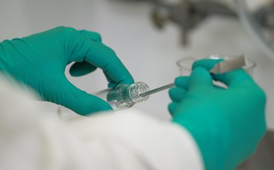 Two hands of an operator, clad in protective gloves, skillfully dispensing a fine white powder from a glass vial using a spatula. Two hands of an operator, clad in protective gloves, skillfully dispensing a fine white powder from a glass vial using a spatula.